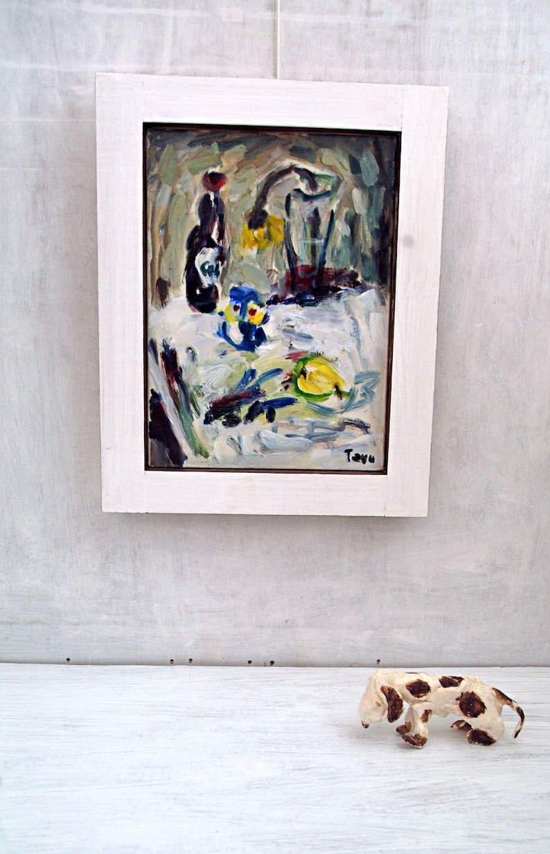May include: A framed painting of a still life with a bottle, a vase, and fruit on a white tablecloth. The painting is in a white frame and has a signature in the bottom right corner.