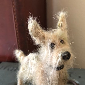 Puede incluir: Figura de perro terrier hecha a mano, de color blanquecino, con nariz y ojos negros. El perro tiene pelo largo y desgre&ntilde;ado y est&aacute; sobre una superficie met&aacute;lica gris. Un objeto decorativo r&uacute;stico y encantador.