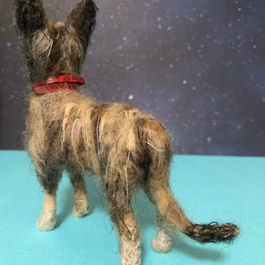 Puede incluir: Figura de perro hecha a mano con pelaje marr&oacute;n y beige, con un collar rojo. El perro est&aacute; visto desde atr&aacute;s, mostrando su cola y patas. El fondo es azul oscuro con motas blancas.