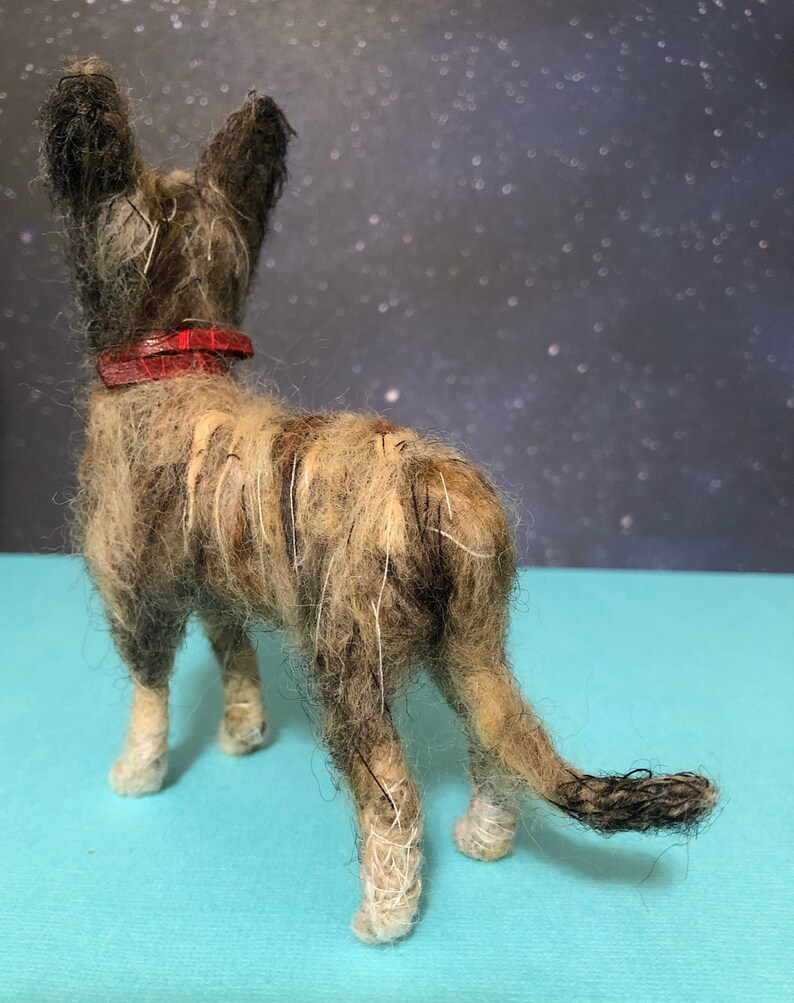 Puede incluir: Figura de perro hecha a mano con pelaje marr&oacute;n y beige, con un collar rojo. El perro est&aacute; visto desde atr&aacute;s, mostrando su cola y patas. El fondo es azul oscuro con motas blancas.