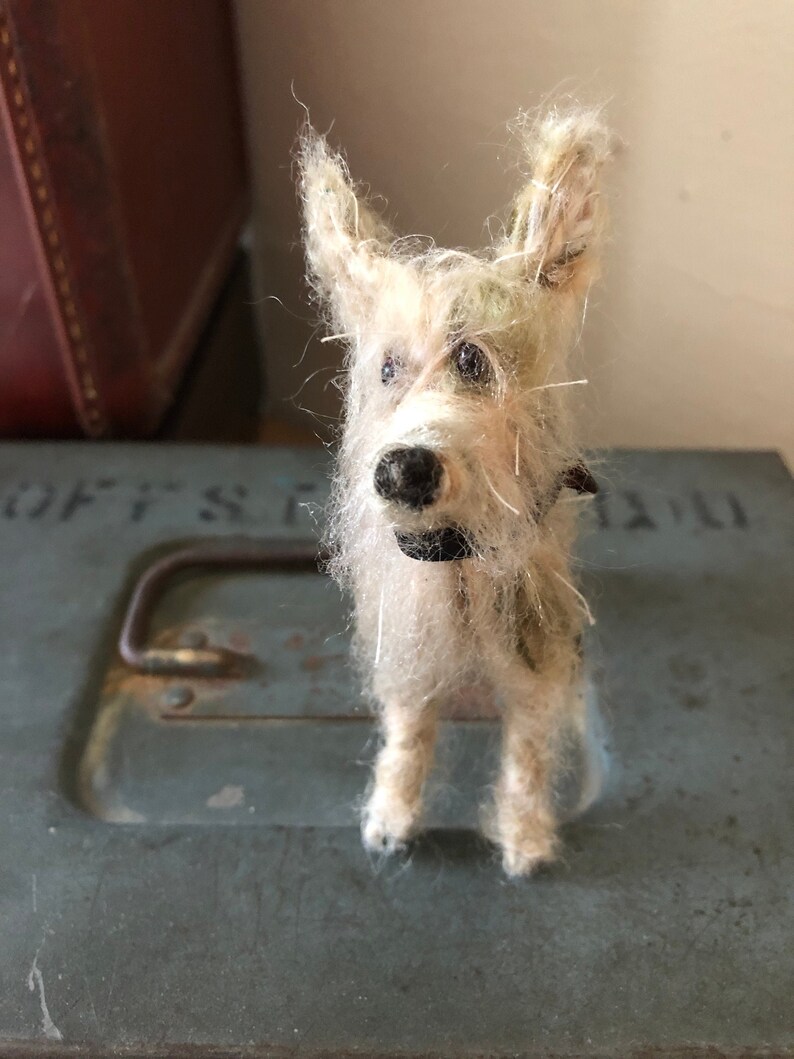 Puede incluir: Figura de perro en miniatura hecha a mano, con un pelaje desgre&ntilde;ado de color claro y ojos y nariz negros. El perro lleva un collar negro y est&aacute; sobre una superficie met&aacute;lica. La figura tiene un estilo caprichoso y de arte popular.