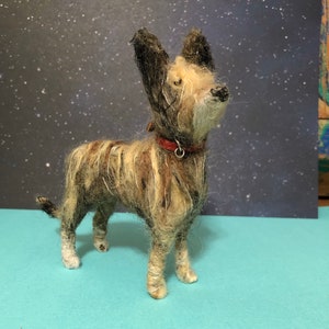 Puede incluir: Figura de perro de fieltro hecha a mano con un collar rojo. El perro tiene un aspecto peludo y texturizado con una mezcla de fibras marrones, beige y blancas. Se apoya sobre cuatro patas, con la cabeza inclinada hacia arriba. El fondo es azul oscuro con puntos blancos.