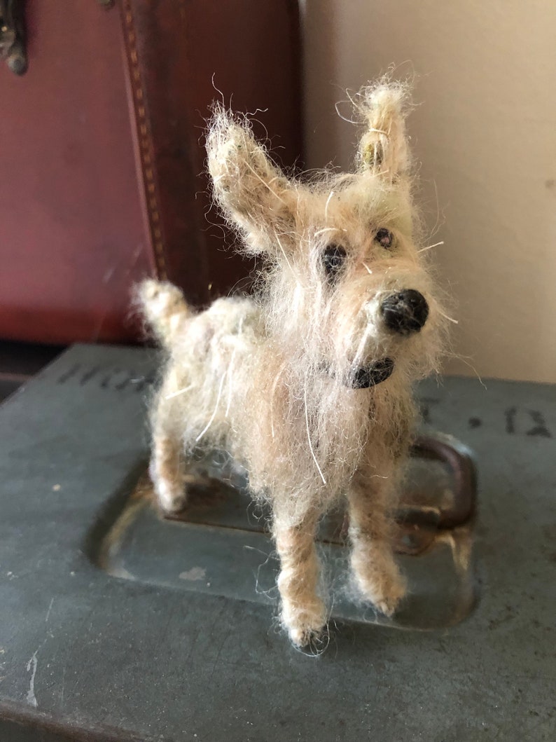 Puede incluir: Figura de perro terrier hecha a mano, de color blanquecino, con nariz y ojos negros. El perro tiene pelo largo y desgre&ntilde;ado y est&aacute; sobre una superficie met&aacute;lica gris. Un objeto decorativo r&uacute;stico y encantador.