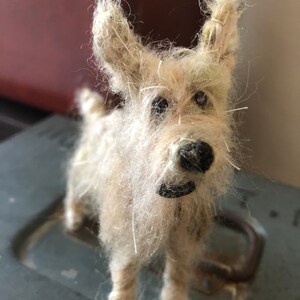 Puede incluir: Una figura de perro en miniatura hecha a mano con un pelaje desgre&ntilde;ado de color claro. El perro tiene una nariz negra y un peque&ntilde;o collar negro. La figura est&aacute; de pie sobre una superficie gris, con un fondo borroso.