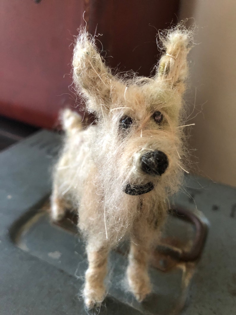 Puede incluir: Una figura de perro en miniatura hecha a mano con un pelaje desgre&ntilde;ado de color claro. El perro tiene una nariz negra y un peque&ntilde;o collar negro. La figura est&aacute; de pie sobre una superficie gris, con un fondo borroso.