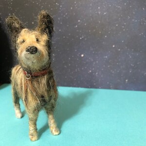 Puede incluir: Figura de perro de fieltro hecha a mano con un collar rojo. El perro tiene un pelaje marr&oacute;n y beige, orejas negras y una nariz negra. Se encuentra sobre una superficie azul claro contra un fondo oscuro con puntos blancos.