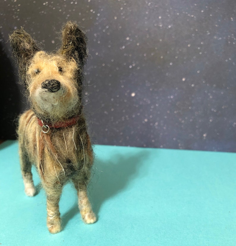 Puede incluir: Figura de perro de fieltro hecha a mano con un collar rojo. El perro tiene un pelaje marr&oacute;n y beige, orejas negras y una nariz negra. Se encuentra sobre una superficie azul claro contra un fondo oscuro con puntos blancos.