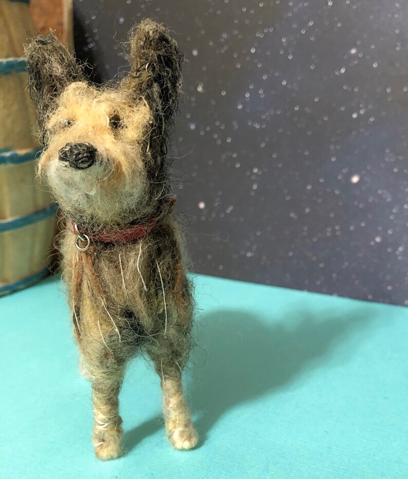 Puede incluir: Figura de perro en miniatura hecha a mano, con pelaje marr&oacute;n y beige, orejas negras y collar rojo. El perro est&aacute; sobre una superficie azul claro. El fondo presenta un dise&ntilde;o de noche estrellada. Aspecto texturizado y detallado.