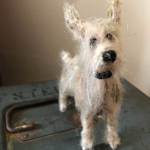 Puede incluir: Figura de perro terrier hecha a mano, de color blanquecino, con nariz y collar negros. El perro tiene un aspecto peludo y texturizado y est&aacute; sobre una superficie met&aacute;lica verde desgastada. Las orejas del perro est&aacute;n erguidas y sus ojos son oscuros.