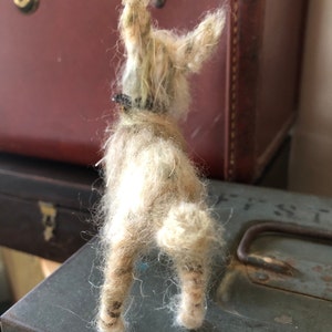 Puede incluir: Figura de perro de fieltro hecha a mano, de color marr&oacute;n claro, vista desde atr&aacute;s. El perro tiene un pelaje suave y texturizado, orejas ca&iacute;das y un peque&ntilde;o collar negro. La figura est&aacute; colocada sobre una superficie met&aacute;lica desgastada, con equipaje vintage en el fondo.