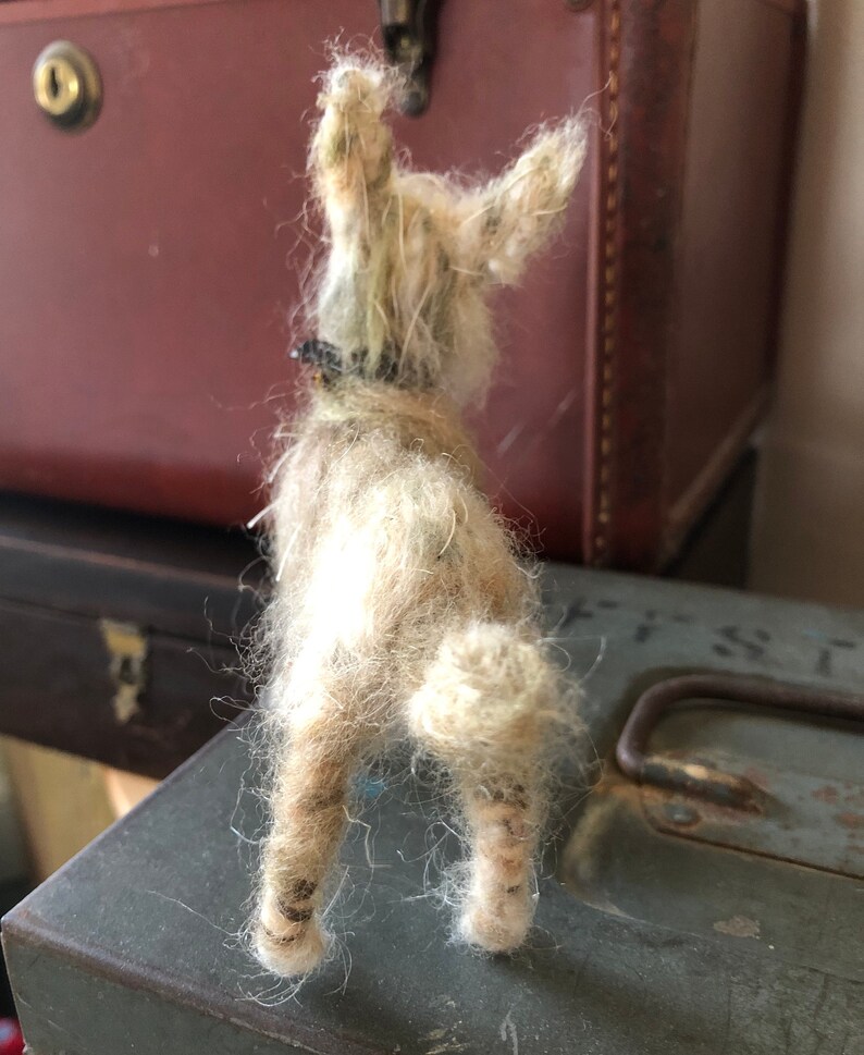 Puede incluir: Figura de perro de fieltro hecha a mano, de color marr&oacute;n claro, vista desde atr&aacute;s. El perro tiene un pelaje suave y texturizado, orejas ca&iacute;das y un peque&ntilde;o collar negro. La figura est&aacute; colocada sobre una superficie met&aacute;lica desgastada, con equipaje vintage en el fondo.