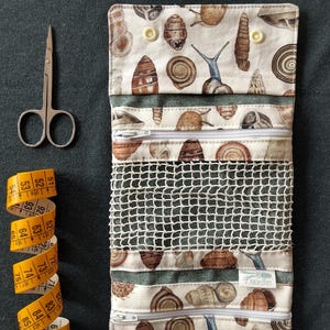 Puede incluir: Un organizador de tela enrollable con un estampado de conchas de caracol en tonos marrón y crema. El organizador incluye dos bolsillos con cremallera, un compartimento de malla y dos cierres a presión. Unas tijeras de metal y una cinta métrica.
