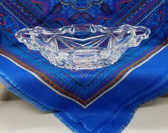 May include: A clear glass bowl with a starburst pattern and two handles. The bowl is sitting on a blue and white paisley patterned fabric.
