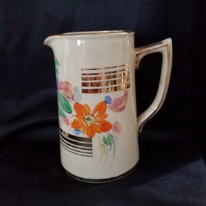 May include: A white ceramic pitcher with a gold rim and handle. The pitcher has a floral design with orange, pink, and green flowers. The background is a cream color with gold stripes.