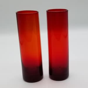 May include: Two red glass shot glasses with a gradient of color from orange to red. The glasses are cylindrical in shape and have a dark red base.