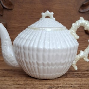May include: A white ceramic teapot with a textured, ribbed body and a coral-like handle. The spout and lid are also white. The teapot is a decorative item, perfect for serving tea. The teapot is approximately 6 inches tall.