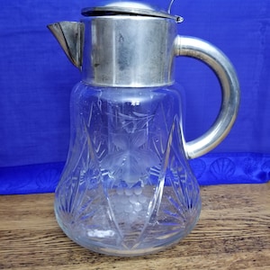 May include: A clear glass pitcher with a silver lid and handle. The pitcher has a cut glass design and a grape motif.
