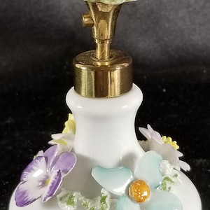 May include: A white porcelain perfume bottle with a gold top and floral accents. The bottle is decorated with purple, blue, and yellow flowers.