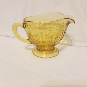 May include: A yellow glass creamer with a floral pattern and a handle. The creamer has a rounded base and a slightly flared lip.