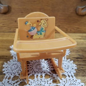 May include: A vintage, orange plastic doll high chair. The chair features a tray and a backrest with a cartoon giraffe and bear design. The chair is approximately 5 inches tall and sits on a white lace doily.