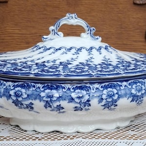 May include: A white and blue ceramic serving dish with a lid. The dish features a floral pattern in blue and white. It has two handles on the sides and a handle on the lid. The dish is likely vintage or antique.