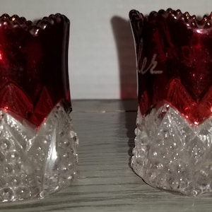 May include: Two small, clear glass cups with a red, glass top. The top of each cup has a white inscription. The cups have a textured, diamond-shaped pattern.
