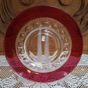 May include: A clear glass plate with a ruby red rim. The plate has a clear center with a design and a ring of small, raised circles. The plate is displayed on a clear plastic stand, showcasing its design and color.