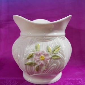 May include: A white ceramic vase with a flared top and a decorative floral design. The vase features a raised pink flower with yellow center and green leaves. The body of the vase has a textured, wood-like pattern.