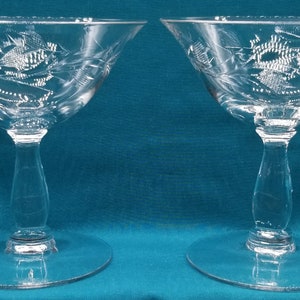 May include: Two clear glass stemmed cocktail glasses with a delicate etched floral design. The glasses are on a teal blue surface.