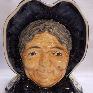 May include: A ceramic figurine of a woman's head wearing a black bonnet with a white trim. The woman has gray hair and a kind smile. The figurine is painted in shades of blue, white, and brown.