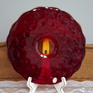 May include: A ruby red glass plate with a textured surface and a scalloped edge. The center features a yellow and orange design. The plate is displayed on a white stand, showcasing its unique design and color.