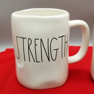 May include: White ceramic mug with the word "STRENGTH" in black lettering. The mug has a rounded shape and a handle. The mug is on a red surface.