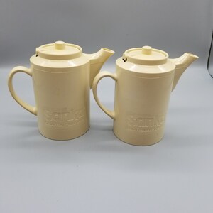 May include: Two cream-colored vintage coffee pots with lids, spouts, and handles. The word "Sanka" is embossed on the front of each pot, along with "Decaffeinated Coffee". The pots are made of a durable plastic material.