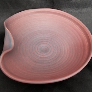 May include: A handcrafted, ceramic plate with a unique, organic shape. The plate features a spiral design in the center and a raised edge. The color is a muted rose with subtle blue accents on the curved side.