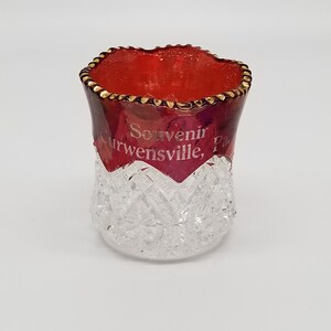 May include: A small, clear glass cup with a red, gold-rimmed top. The cup has a diamond-shaped pattern and the text "Souvenir Curwensville, Pa." is printed on the red rim.