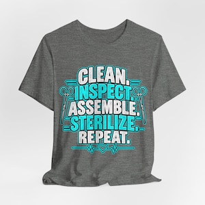 May include: A dark heather grey short-sleeve t-shirt featuring a graphic print in white and bright blue. The text reads CLEAN, INSPECT, ASSEMBLE, STERILIZE, REPEAT, flanked by two pairs of scissors and a heartbeat line with a small heart symbol.