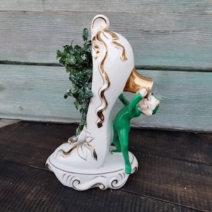 May include: A vintage ceramic shoe-shaped vase, white with gold accents, featuring a green elf figure and green foliage. The shoe has a gold heel and decorative gold trim. The base has a decorative swirl design.