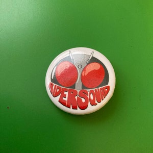 Könnte beinhalten: Ein runder, weißer Knopf mit einem Cartoon-Design. Es zeigt zwei große rote Augen und den Text "RIDERSQUAD" in Rot. Der Knopf befindet sich vor einem grünen Hintergrund.