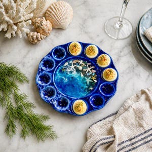 May include: A blue ceramic deviled egg serving platter with a coastal design. The center features a blue ocean scene with starfish and shells. The platter holds deviled eggs, each topped with paprika. Seashells and coral are arranged around the platter.