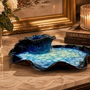 May include: A decorative glass serving dish and small bowl set. The dish is a deep blue with a wavy rim and a light blue and white interior. The bowl is a darker blue with a similar wavy design. The set is on a marble surface.