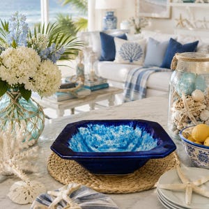 May include: A coastal-themed interior scene featuring a blue and white color palette. A square, dark blue ceramic bowl with a textured, light blue interior sits on a woven placemat. Decorative elements include seashells, starfish, and a glass jar filled with beach-themed items.