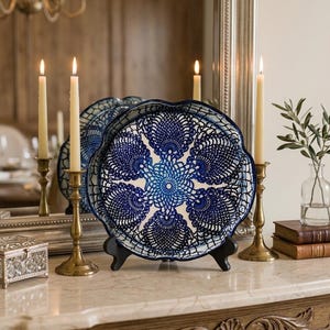 May include: A decorative blue and white plate with a detailed lace-like design, displayed on a black stand. The plate is set on a marble-topped table, with lit candles in brass holders and a mirror in the background.