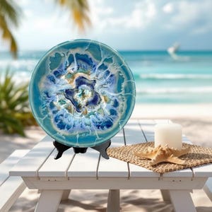 May include: A decorative ceramic plate with a blue and white abstract design, displayed on a black stand. The plate is on a white picnic table with a candle and starfish, with a beach and ocean in the background.
