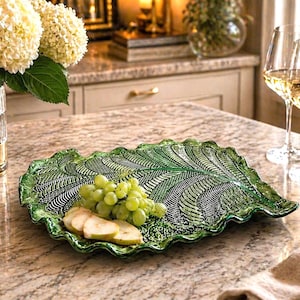 May include: A green leaf-shaped serving platter with a textured design, holding green grapes and sliced pears. The platter is on a granite countertop, with wine glasses and a bottle of wine in the background. Decorative flowers are in a vase.
