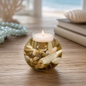 May include: A round glass candle holder filled with seashells and glitter, with a lit candle inside. The candle holder sits on a wooden surface, with a blurred beach scene in the background. The candle flame is bright.