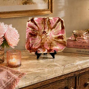 May include: A decorative square plate with a pink and brown floral design, displayed on a black stand. The plate is set on a marble surface, alongside a lit candle, a vase of pink flowers, and books.