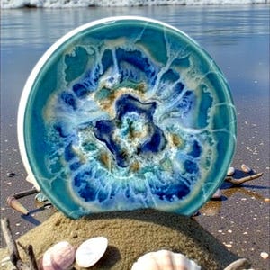 May include: A round, ceramic decorative plate with a white rim. The plate features a swirling design in shades of blue, teal, and beige, resembling an aerial view of the ocean. The plate is resting on a small sand dune with seashells and driftwood.