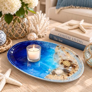 May include: An oval resin tray featuring a blue and white ocean wave design with a sandy beach section. The tray is decorated with a small turtle figurine, seashells, and starfish, and holds a clear glass votive candle holder with a white candle.