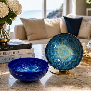May include: A decorative arrangement featuring a blue bowl and a matching decorative sphere on a gold stand. A stack of books and a vase of white flowers are also present. The sphere has a unique, textured design.
