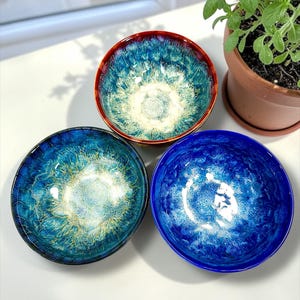 May include: Three handmade ceramic bowls with a glossy finish. The bowls are various shades of blue, with a red rim on one. The bowls have a unique, textured appearance, and are arranged on a white surface.